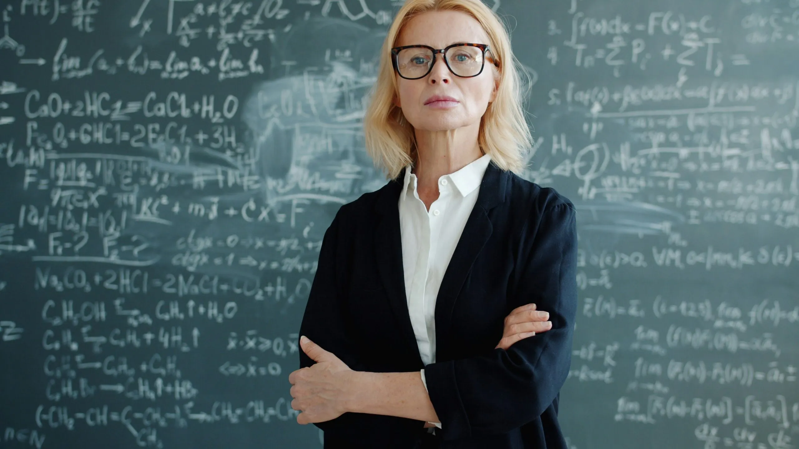 A woman in glasses stands before a chalkboard with equations.