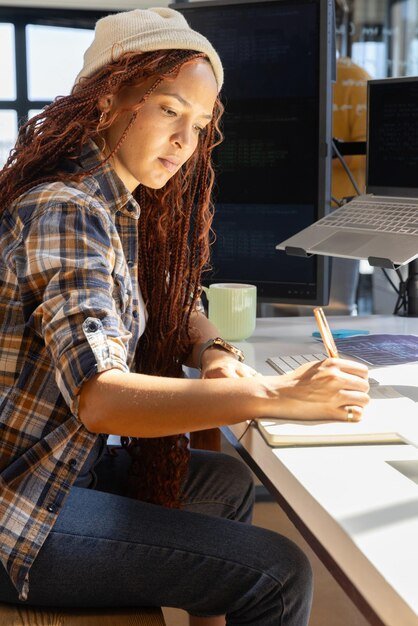 African American woman software developer sitting at office desk taking notes beside dual monitors