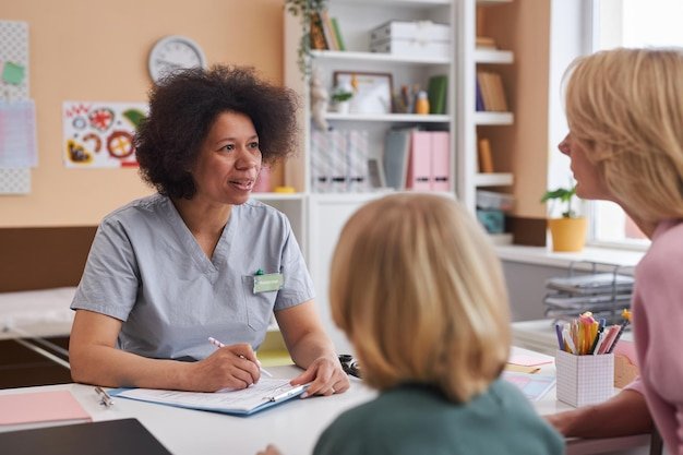 Black young woman as pediatrician talking to mother with child in clinic