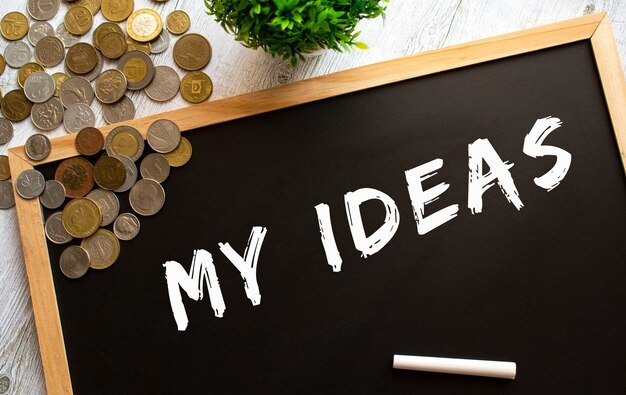 Blackboard with the text MY IDEAS and metal coins on a gray wooden table. Financial concept.