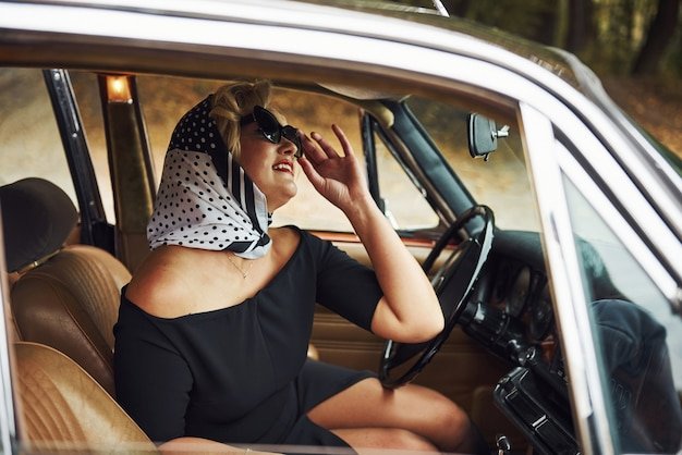 Blonde woman in sunglasses and in black dress sits in old vintage classic car.