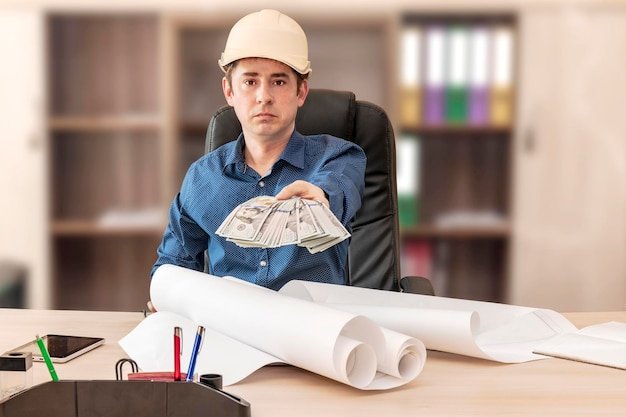 The boss in the construction helmet at the desk with the design documents is holding out a bundle of dollars Successful completion of the project Builder engineer head foreman