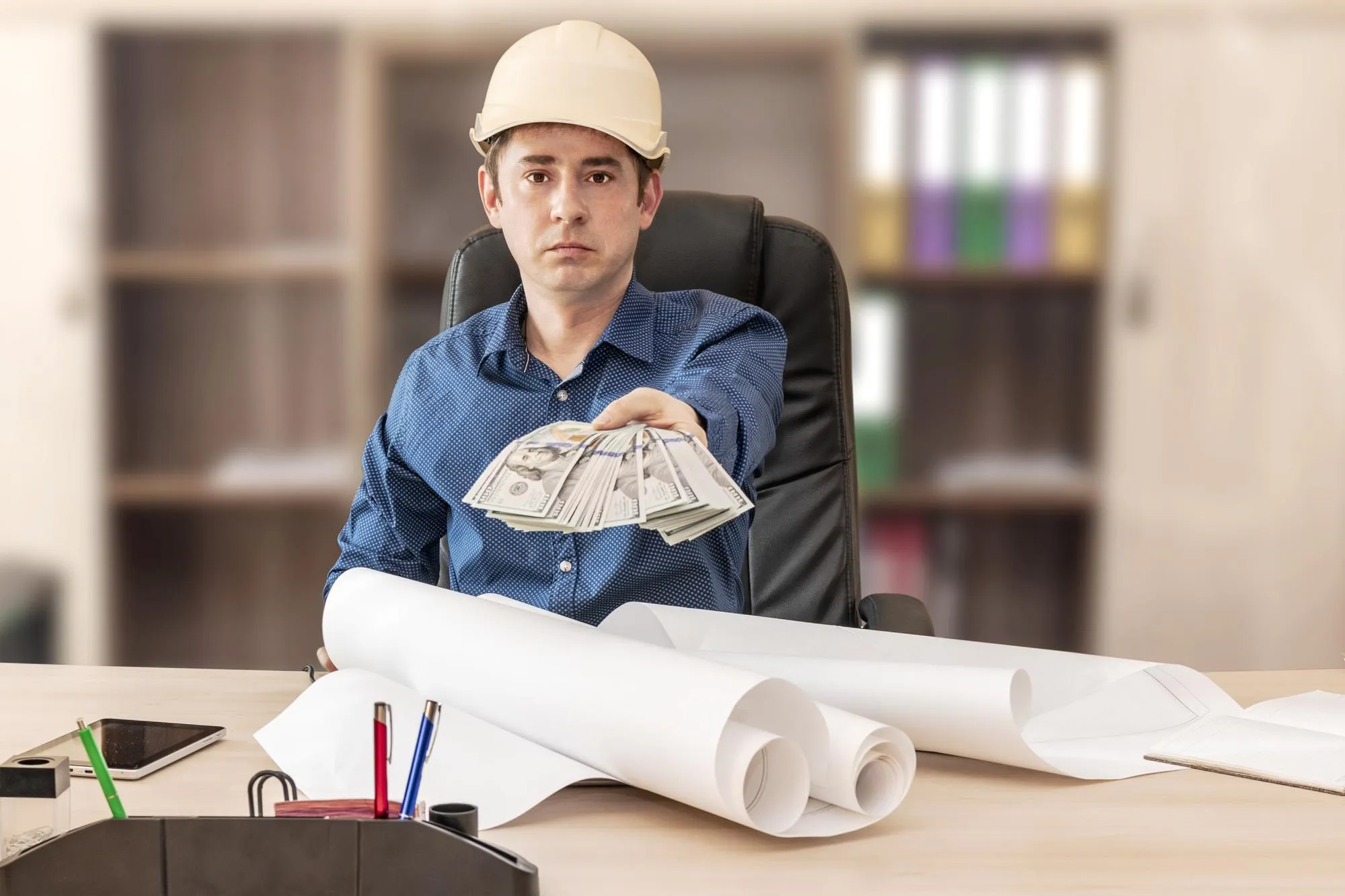 The boss in the construction helmet at the desk with the design documents is holding out a bundle of dollars Successful completion of the project Builder engineer head foreman