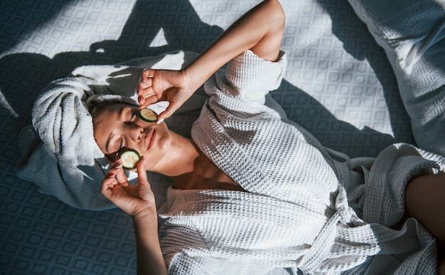 Cheerful mood. Young positive woman with towel on head lying on the bed with cucumber.