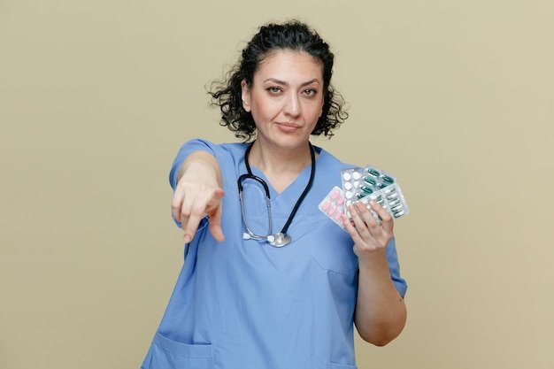 Confident middleaged female doctor wearing uniform and stethoscope around neck showing packs of pills looking and pointing at camera isolated on olive background