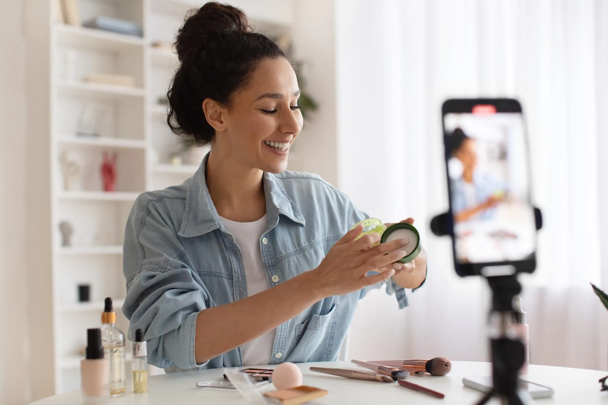 Female blogger showing skincare product to cellphone making video indoor
