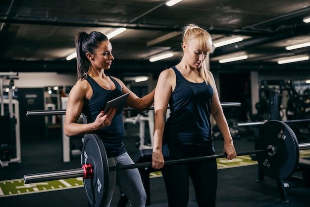 A female coach is training female bodybuilder in a gym wile tracking progress on a tablet