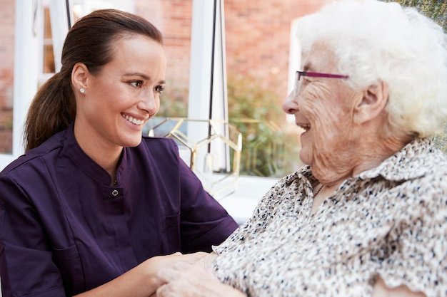 Female Resident Sitting In Chair And Talking With Carer In Retirement Home