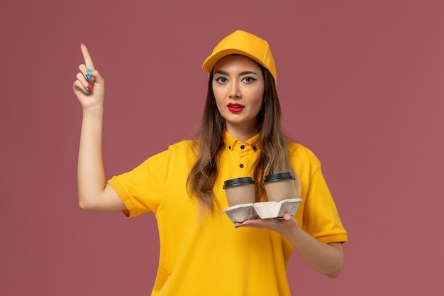 Front view of female courier in yellow uniform and cap holding delivery coffee cups on the pink wall