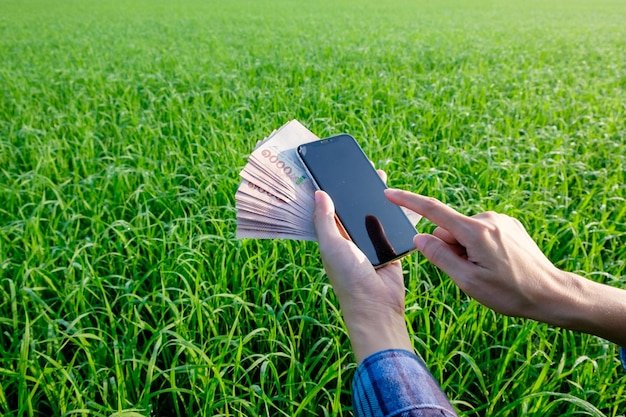 A hand holding a smartphone on grass in a park with money banknote surrounded by nature and technology