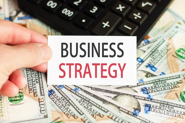 A hand holds a sign with the inscription - BUSINESS STRATEGY on the wall of banknotes and a calculator on the table. Finance and economics concept.