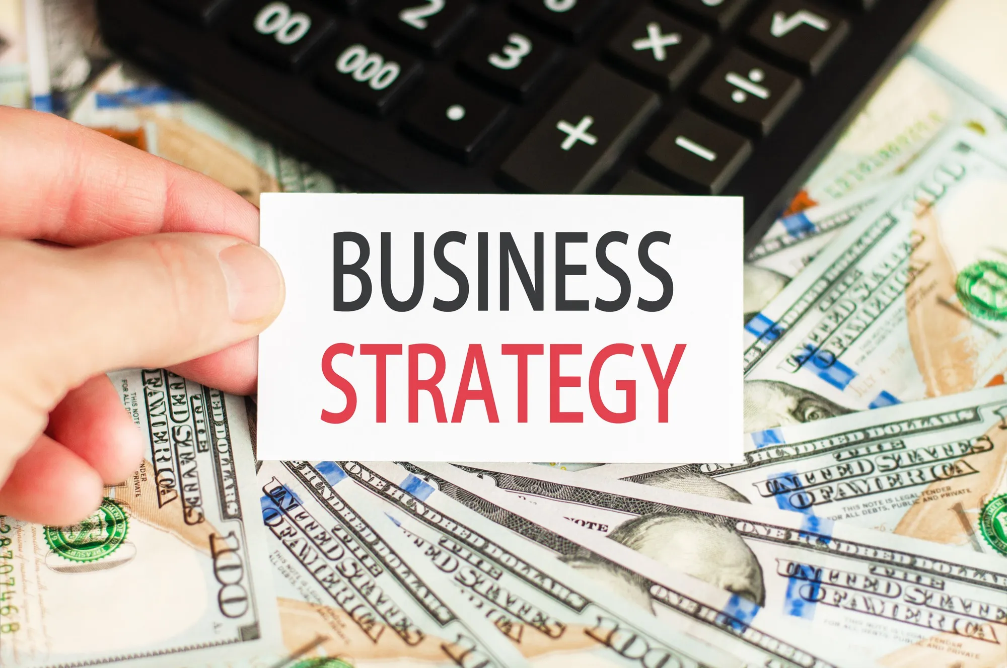 A hand holds a sign with the inscription - BUSINESS STRATEGY on the wall of banknotes and a calculator on the table. Finance and economics concept.