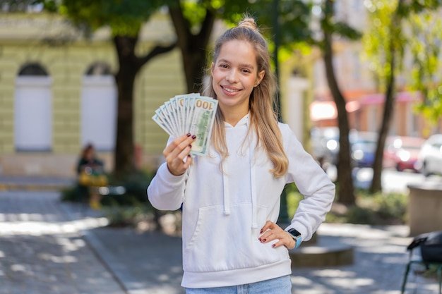 Happy Caucasian young woman girl holding fan of dollars showing thumbs up gesture on city street