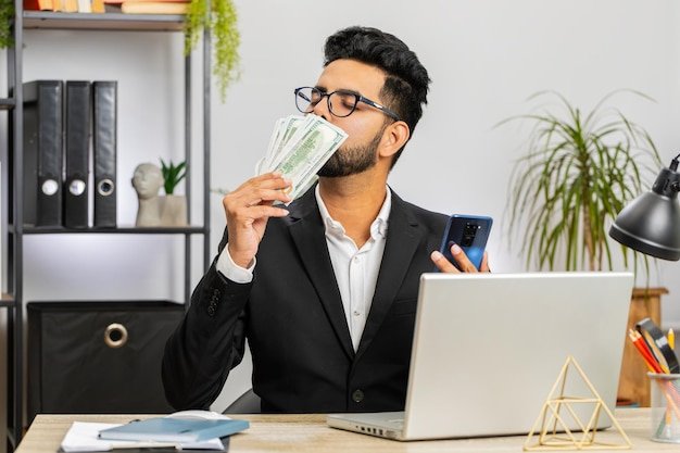 Happy rich Indian businessman counting money cash use smartphone app earning profit success contract