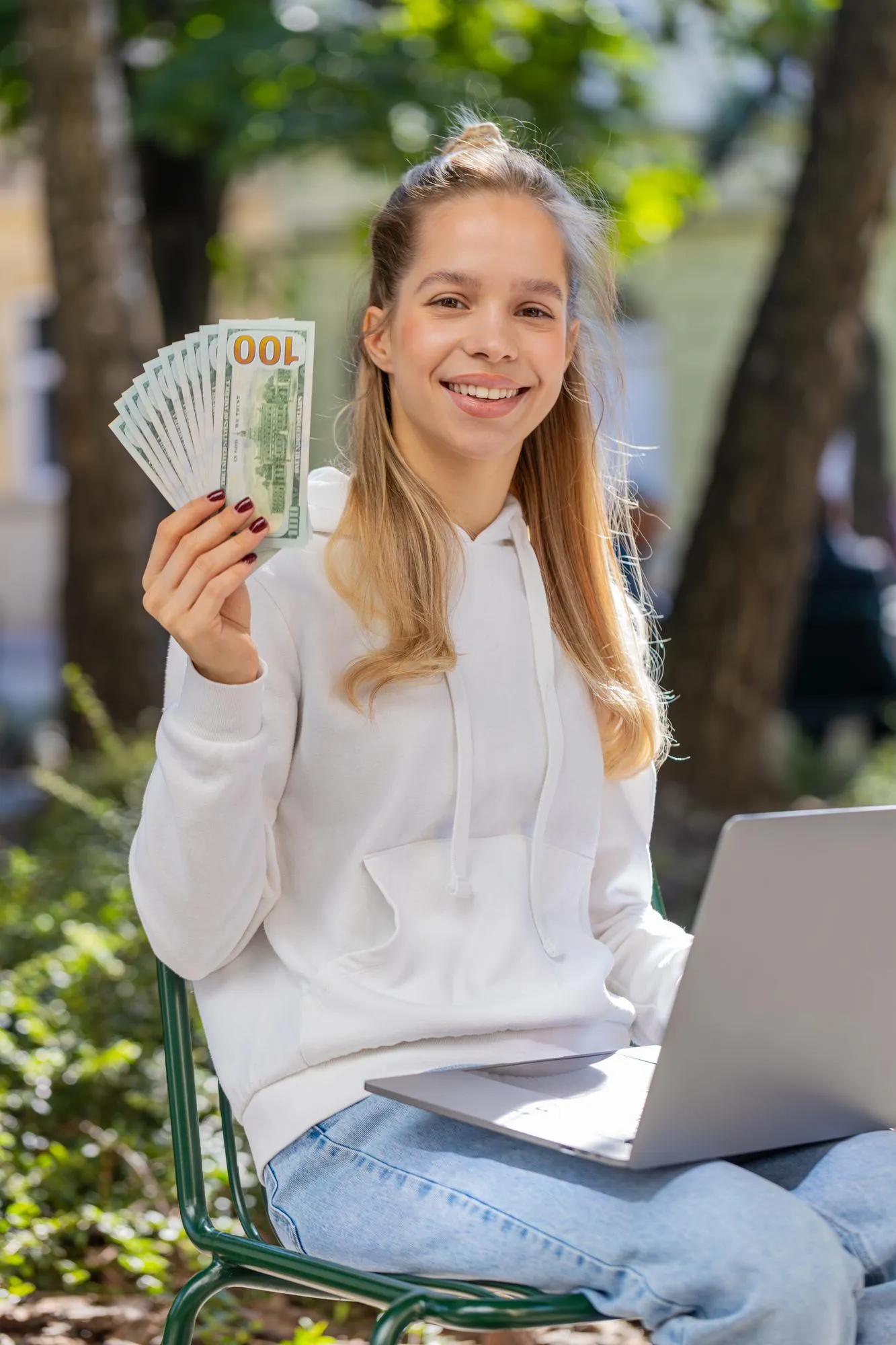 Happy rich woman using laptop winner victory holding cash money in dollar banknotes sitting on chair