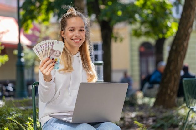 Happy rich woman using laptop winner victory holding cash money in Ukrainian hryvnias UAH banknotes
