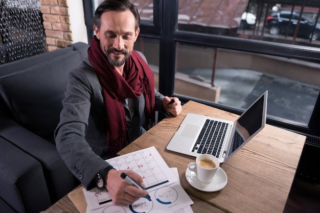 I enjoy my job. Positive handsome delighted man holding a highlighter and writing with it while working in the cafe