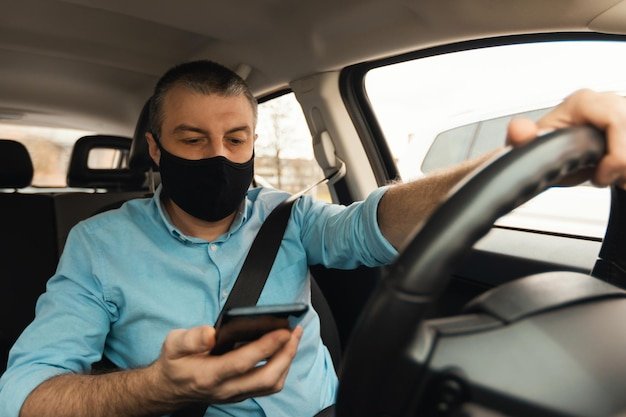 Man driving car using smartphone wearing face mask in automobile