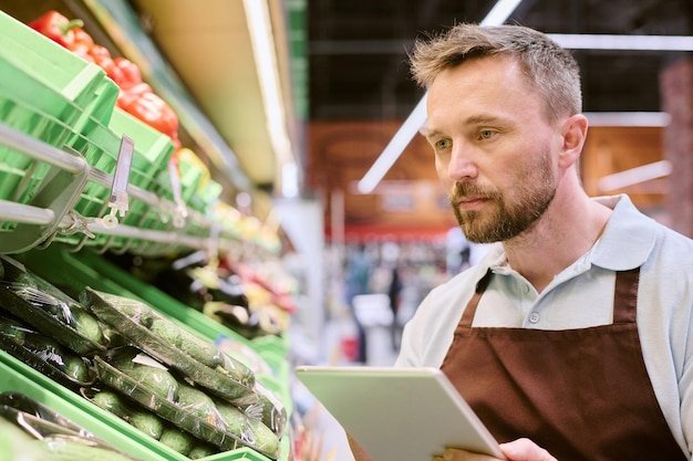 Man in store wearing apron holding tablet while checking produce inventory background shows various