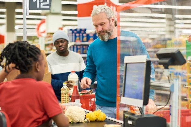 Mature bearded man standing by counter line in front of black female cashier