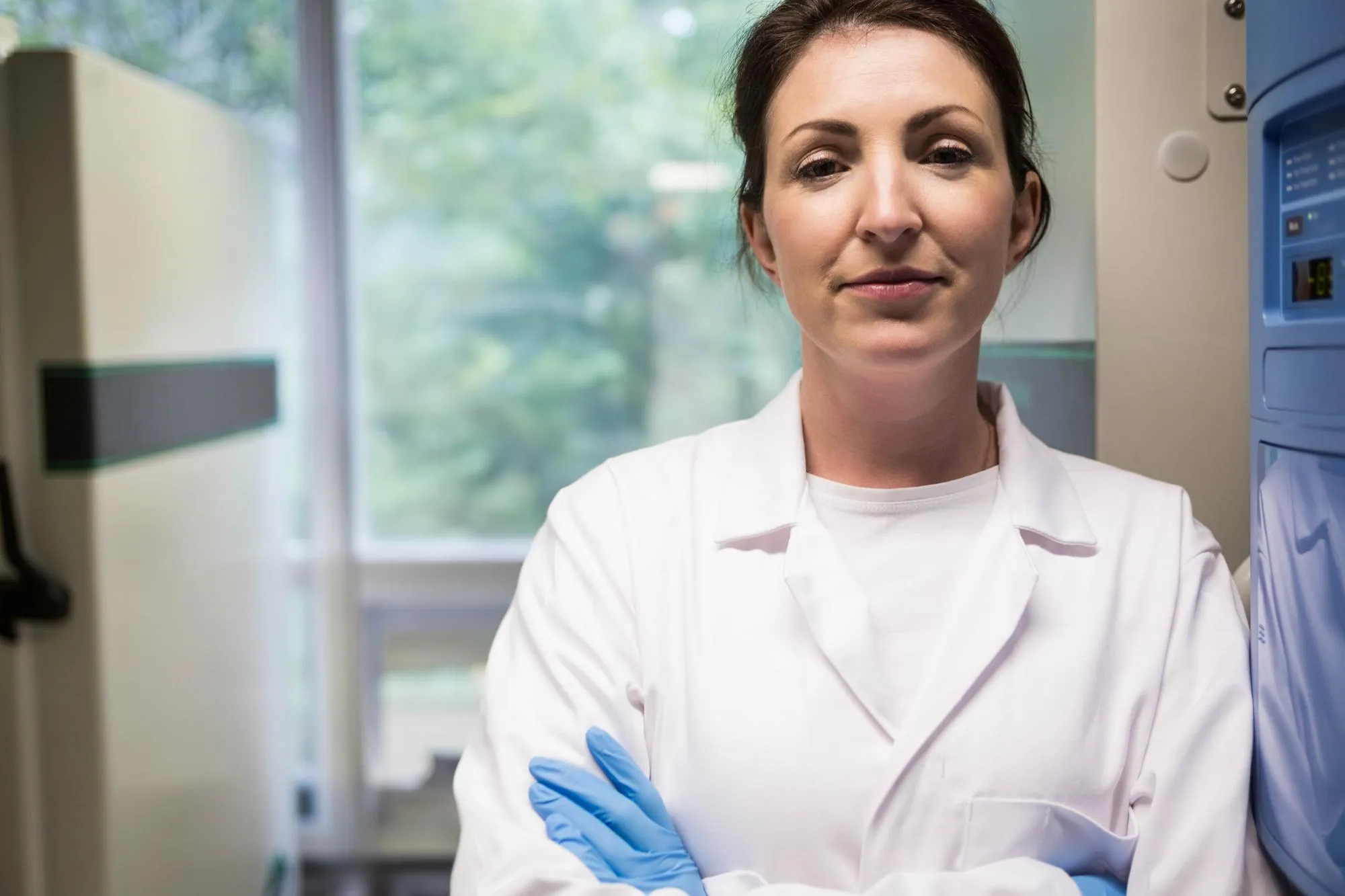 Mid adult female scientist wearing lab coat and blue gloves standing beside freezer in laboratory