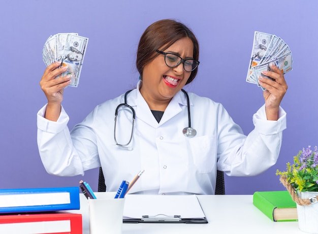 Middle age woman doctor in white coat with stethoscope holding cash happy and excited rejoicing her success sitting at the table over blue background