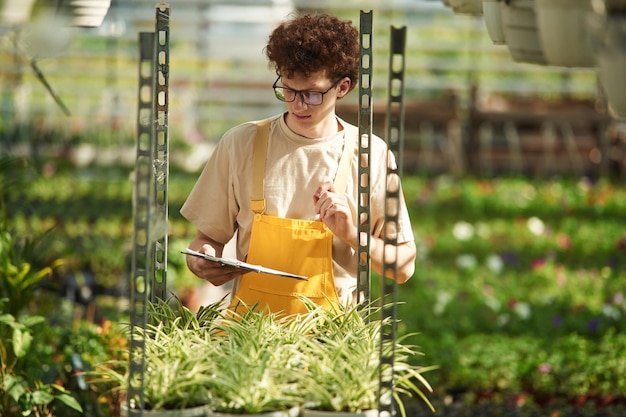 Notepad in hands Young man with curly hair and in glasses is in greenhouse