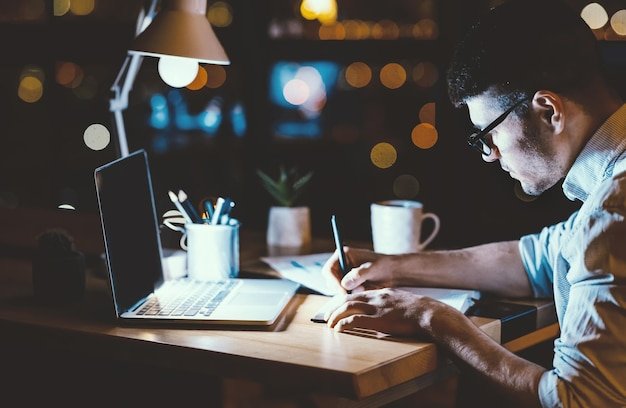 Office manager guy working overnight sitting at laptop computer in modern office low light