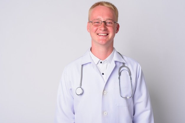 Portrait of young man doctor with blond hair wearing eyeglasses against white wall