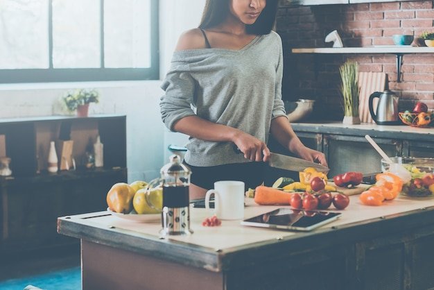 Preparing healthy food. Close-up of beautiful young mixed race woman cutting vegetables for salad while standing in kitchen at home