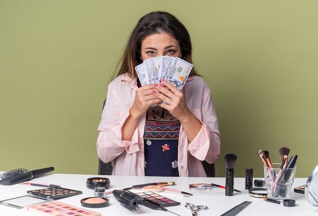 Pretty young brunette girl sitting at table with makeup tools holding money 