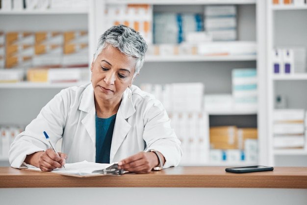 Senior woman pharmacist writing and paper data of pharmacy stock or insurance documents Healthcare doctor and elderly female wellness consultant with store logistics research about shop products
