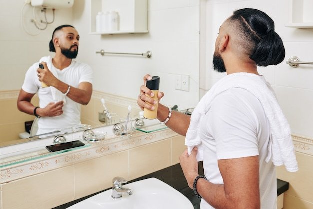 Serious man looking at bathroom mirror and spraying perfume