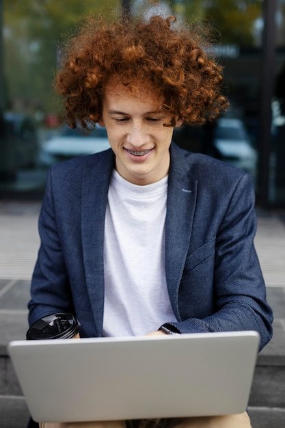 Smiling attractive man using laptop outdoors Successful programmer sitting near modern office