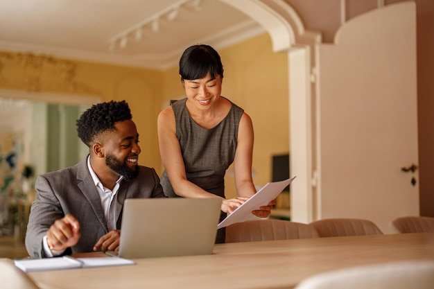 Smiling businesswoman showing reports to male colleague working over laptop at desk in modern office