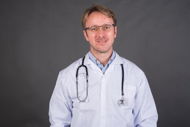 Smiling male doctor with stethoscope over neck in medical coat against gray background