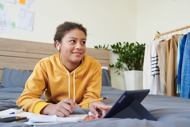 Smiling pretty Mixed-race teenage girl in wireless headphones lying on bed with gray linen and using tablet while performing task
