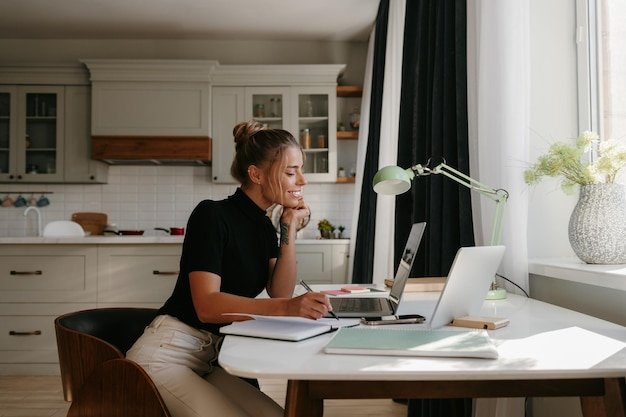 Smiling young woman making notes and looking at the laptop while working at home