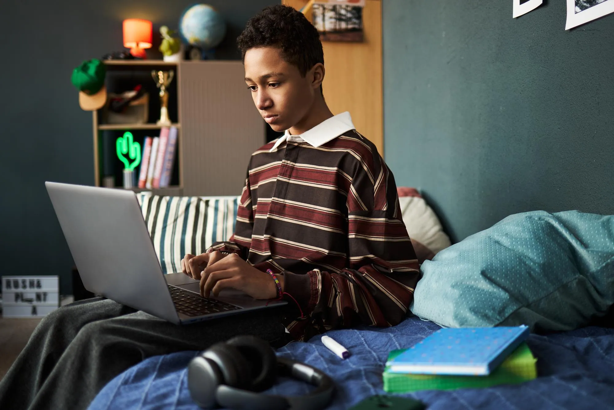 Teenage black boy sitting on bed using laptop for studying at home