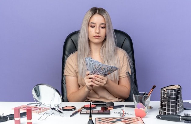 Thinking young beautiful girl sits at table with makeup tools holding and looking at cash isolated on blue background