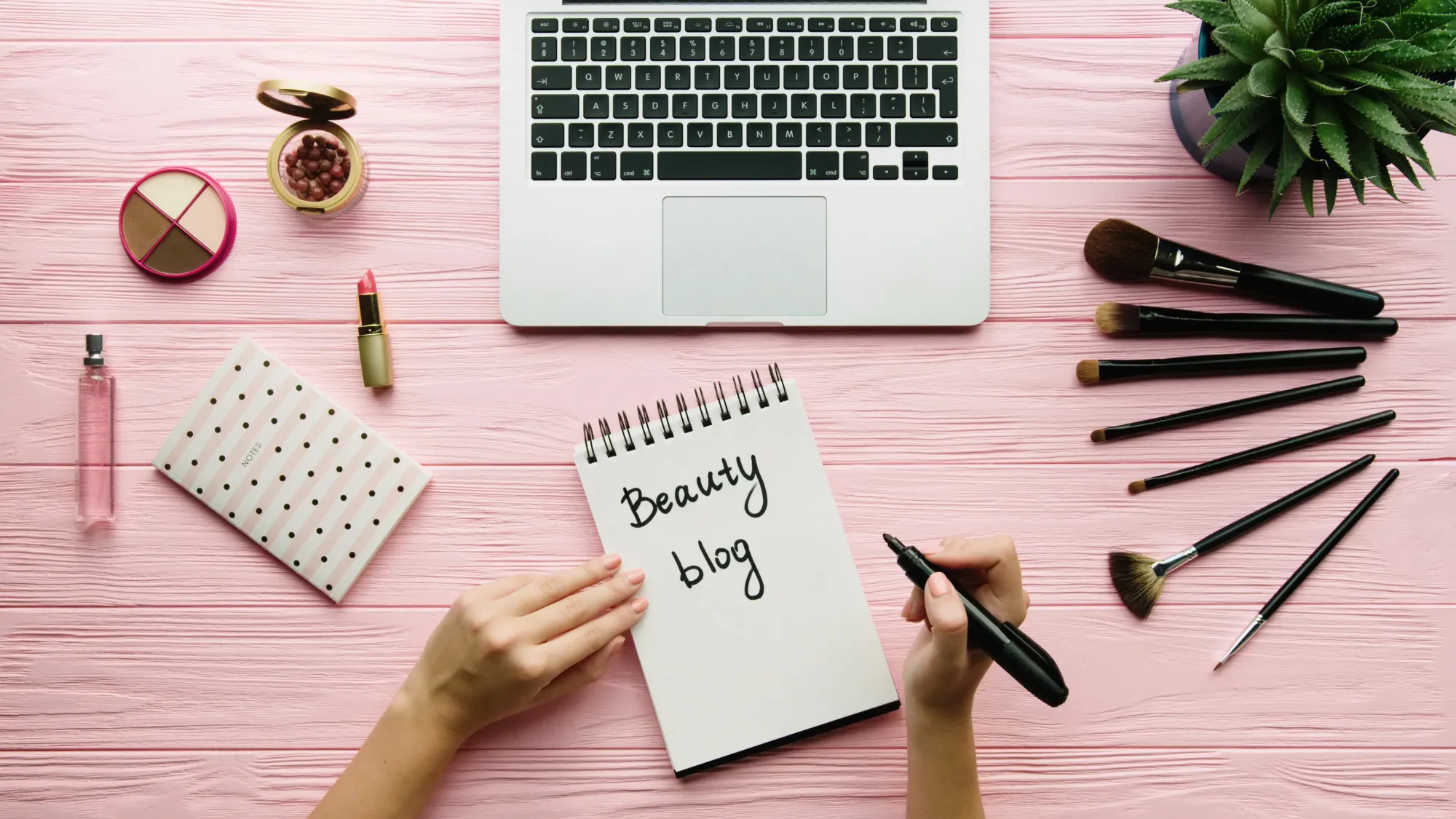 Top view of creative decorated composition with cosmetics, makeup tools, accessory and woman hands writing on notebook on color surface. Beauty, fashion and shopping concept.