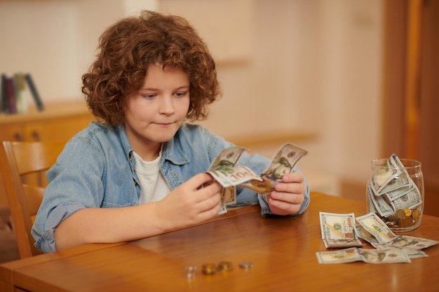 A wavy-haired boy in a denim shirt counting his savings