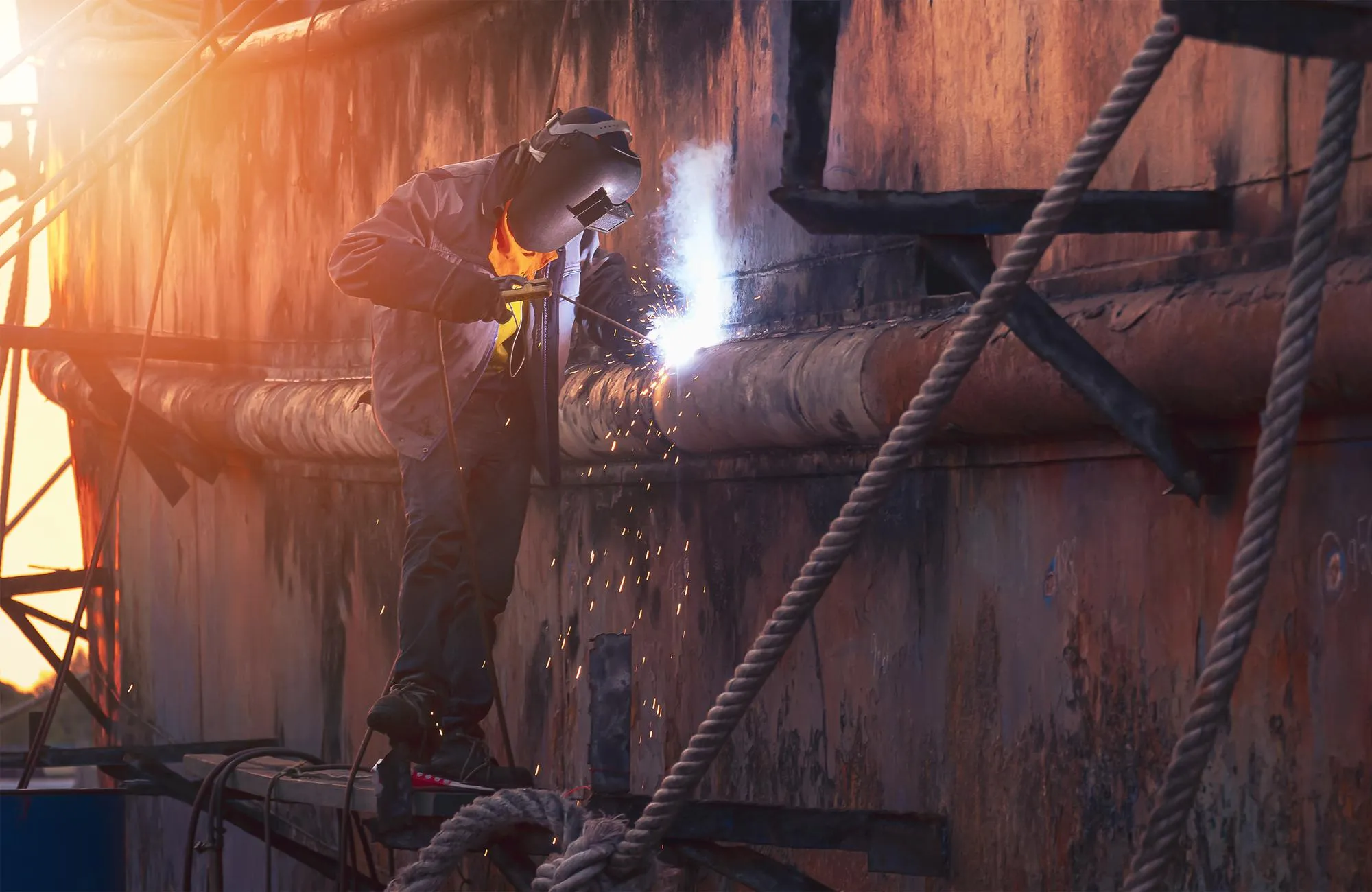 Welder on platform is welding the old vessel hull in shipyard area at sunset time