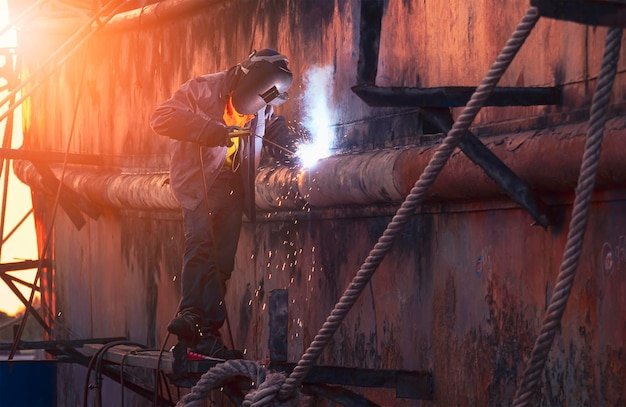 Welder on platform is welding the old vessel hull in shipyard area at sunset time
