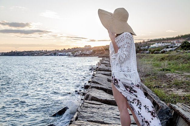 Woman by the sea at sunset back view. Dressed in a long skirt and hat.