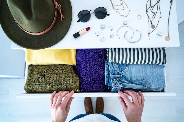 Woman choosing outfit from drawer dresser with stylish casual trendy clothes, home stuff and female accessories. Top view.