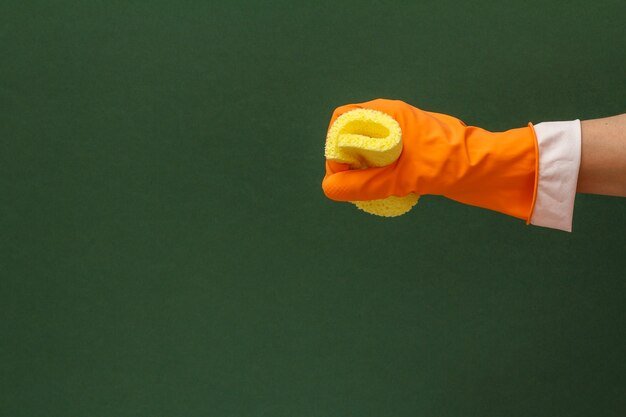 Woman's hand in orange protective glove with yellow sponge in a green background. Washing and cleaning concept.