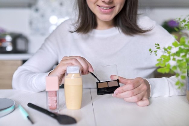Young attractive woman doing makeup, female sitting at home at table in kitchen with cosmetic products mirror brush, shows eyebrow shadow makeup. Beauty, skin care, fashion, make up concept