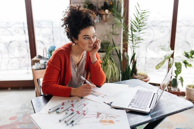 Young beautiful woman with dark curly hair sitting at the table with laptop and fashion sketches dreamily looking aside while spending time in modern cozy workshop with big windows