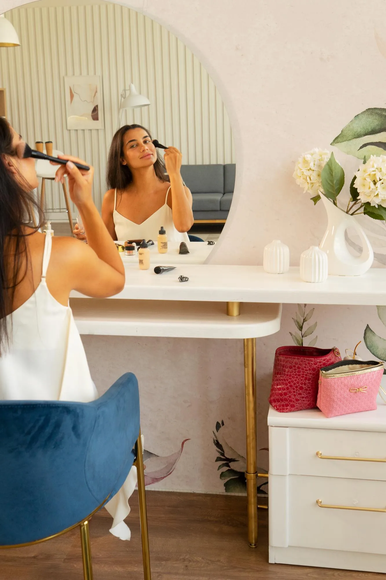 Young beautiful woman with long straight dark hair and fresh clean skin doing morning makeup routine with brush while sitting in front of mirror table at home Beauty and facial cosmetics concept